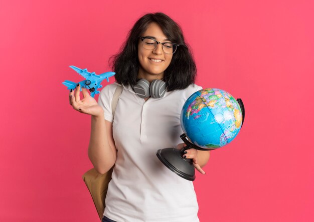 Das junge erfreute hübsche kaukasische Schulmädchen mit Kopfhörern am Hals, das Brille und Rückentasche trägt, hält Spielzeugflugzeug und Globus auf Rosa mit Kopienraum