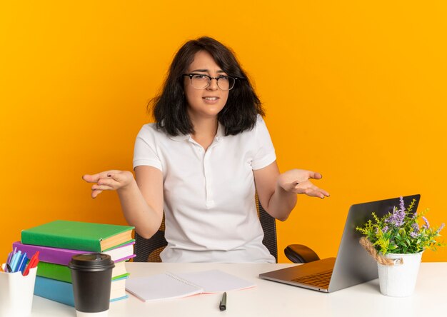 Das junge genervte hübsche kaukasische Schulmädchen, das eine Brille trägt, sitzt mit Schulwerkzeugen am Schreibtisch und hält die Hände auf Orange mit Kopierraum offen