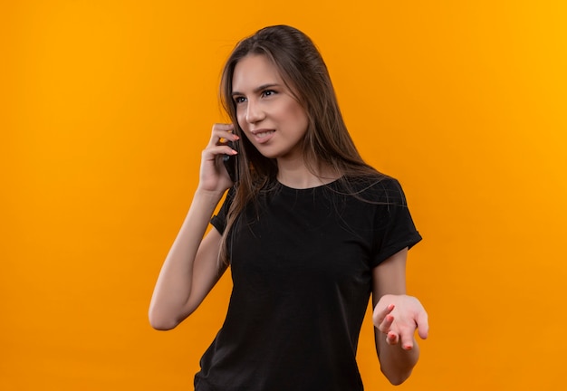 Das junge kaukasische Mädchen der Seite, das schwarzes T-Shirt trägt, spricht am Telefon auf lokalisiertem orange Hintergrund