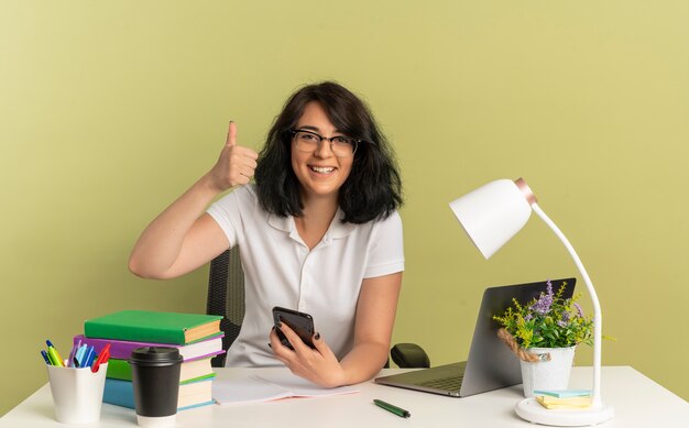 Das junge lächelnde hübsche kaukasische Schulmädchen, das eine Brille trägt, sitzt am Schreibtisch mit den Schulwerkzeug-Daumen hoch und hält Telefon lokalisiert auf Grünfläche mit Kopienraum