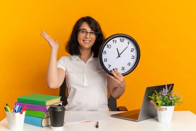 Das junge lächelnde hübsche kaukasische Schulmädchen, das eine Brille trägt, sitzt am Schreibtisch mit Schulwerkzeugen hält Uhr und hebt Hand auf lokalisiertem orange Raum mit Kopienraum hoch