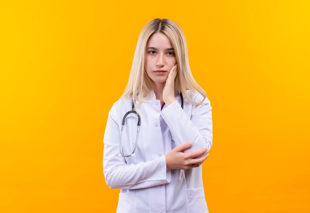 Das junge Mädchen des Doktors, das Stethoskop im medizinischen Kleid trägt, legte ihre Hand auf Wange auf isolierte gelbe Wand