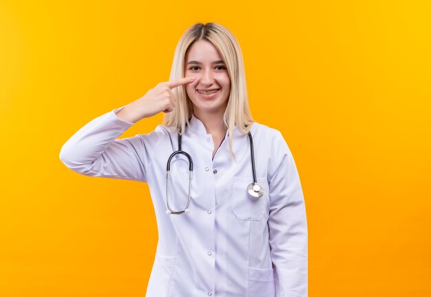 Das junge Mädchen des lächelnden Doktors, das Stethoskop im medizinischen Kleid und in den Zahnspangen trägt, zeigt auf lokalisierten gelben Hintergrund