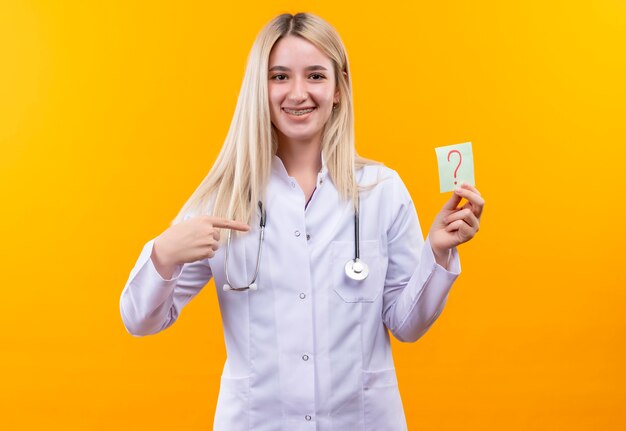 Das junge Mädchen des lächelnden Doktors, das Stethoskop im medizinischen Kleid und in der Zahnspange trägt, zeigt auf Papier-Fragezeichen auf ihrer Hand auf lokalisiertem gelbem Hintergrund