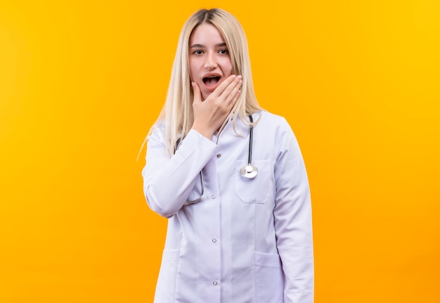 Kostenloses Foto das junge mädchen des traurigen doktors, das stethoskop im medizinischen kleid trägt, legte seine hand auf schmerzenden zahn auf lokalisiertem gelbem hintergrund