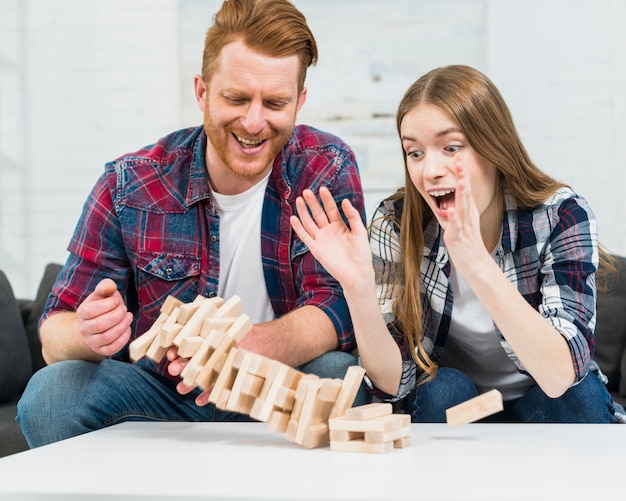 Das junge Paar, das Jenga-Turm betrachtet, bricht auf weißer Tischplatte zusammen