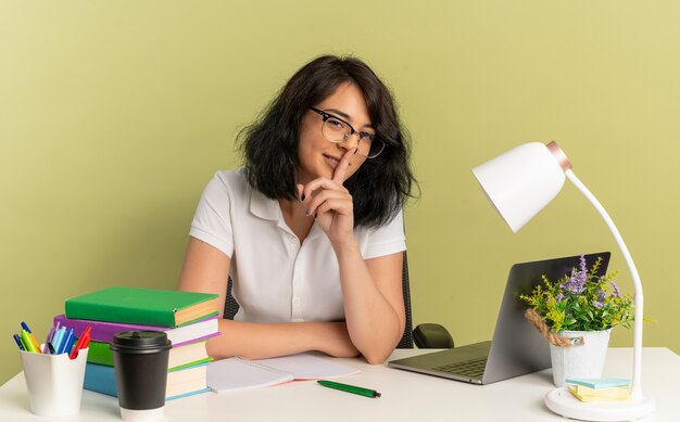 Das junge selbstbewusste hübsche kaukasische Schulmädchen, das eine Brille trägt, sitzt am Schreibtisch mit den Gesten der Schulwerkzeuge