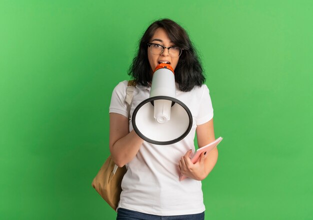 Das junge überraschte hübsche kaukasische Schulmädchen, das Brille und Rückentasche trägt, hält Lautsprecher, der Notizbuch auf Grün mit Kopienraum hält