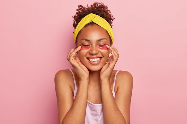 Das junge weibliche Afro-Model trägt Kollagenpads unter den Augen auf, genießt eine feuchtigkeitsspendende Behandlung, lächelt breit, zeigt, dass weiße Zähne frische, gesunde Haut haben und ein gelbes Stirnband tragen