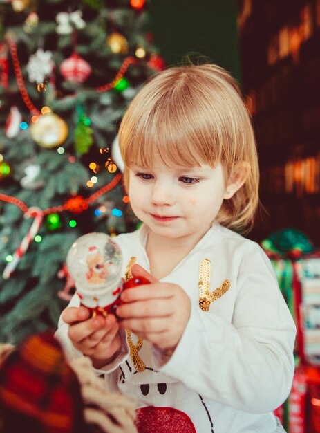 Das kleine Mädchen hält ein Spielzeug und steht in der Nähe von Weihnachtsbaum