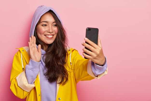 Das Lächeln des angenehm aussehenden asiatischen Mädchens winkt mit der Handfläche und begrüßt die Kamera des modernen Smartphones, tätigt einen Videoanruf, hat lange dunkle Haare, trägt ein lila Sweatshirt und einen gelben Regenmantel und posiert im Innenbereich.