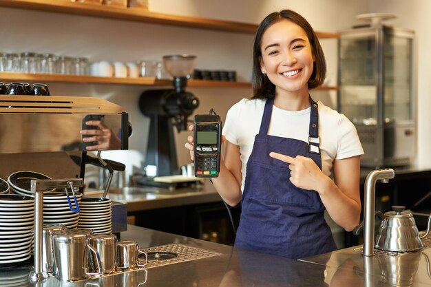 Das lächelnde asiatische Barista-Mädchen trägt eine Schürze und zeigt einen Kreditkartenautomaten für die Zahlungsabwicklung