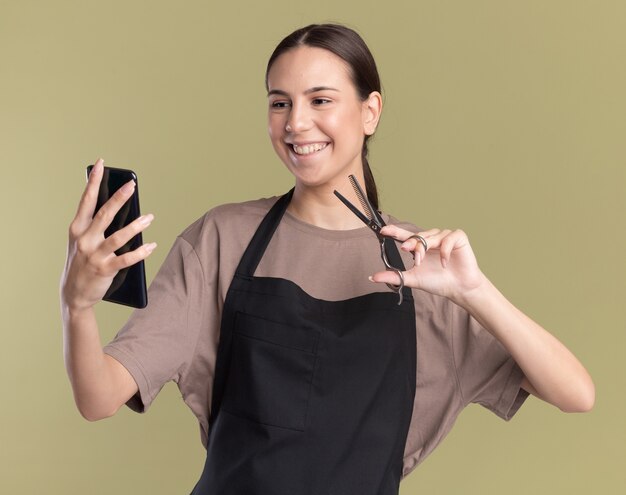 Das lächelnde junge brünette Friseurmädchen in der Uniform hält eine haarausdünnende Schere und schaut auf das Telefon auf Olivgrün