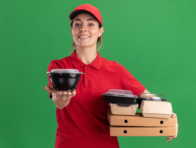 Das lächelnde junge hübsche Liefermädchen in der Uniform hält Papiernahrungsmittelpakete auf Pizzaschachteln und Lebensmittelbehälter auf Grün