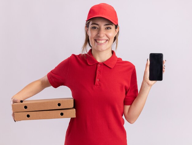 Das lächelnde junge hübsche Liefermädchen in der Uniform hält Pizzaschachteln und Telefon auf Weiß