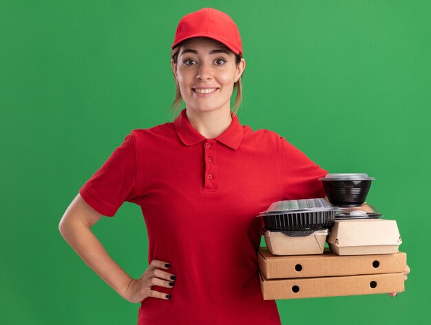 Das lächelnde junge hübsche Liefermädchen in der Uniform legt die Hand auf die Taille und hält Lebensmittelverpackungen aus Papier