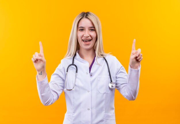 Das lächelnde junge Mädchen des Doktors, das Stethoskop im medizinischen Kleid und in der Zahnspange trägt, zeigt auf lokalisierten gelben Hintergrund