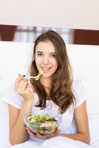 Das lächelnde junge Mädchen isst ihr Gemüsesalatfrühstück im Bett