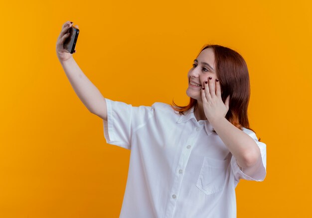 Das lächelnde junge rothaarige Mädchen nimmt ein Selfie und legt Hand auf Wange auf Gelb