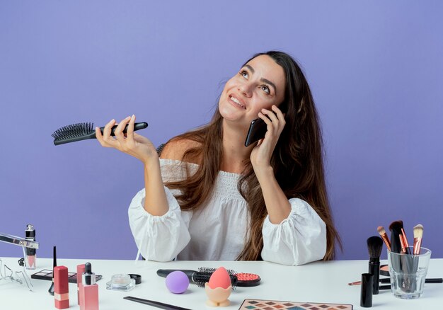 Das lächelnde schöne Mädchen sitzt am Tisch mit Make-up-Werkzeugen und hält den Haarkamm, der am Telefon spricht und isoliert auf lila Wand schaut