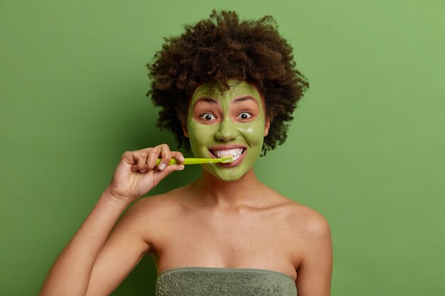 Das lockige junge Mädchen mit Afro-Haar putzt die Zähne und unterzieht sich täglichen Hygieneroutinen. Es wird eine Gesichtsmaske für gesunde Haut angewendet, die in ein Handtuch gewickelt ist, das über einer leuchtend grünen Wand isoliert ist