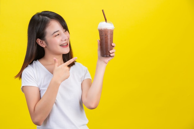 Das Mädchen trinkt kaltes Wasser aus Kakao aus einem durchsichtigen Plastikglas auf gelbem Grund.