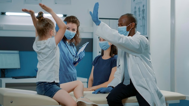 Das medizinische Team gibt dem kleinen Kind nach erfolgreicher Untersuchung High Five und feiert das Ende des Termins. Arzthelferin und Kinderarzt im Gespräch mit fröhlichem Mädchen beim Kontrollbesuch.