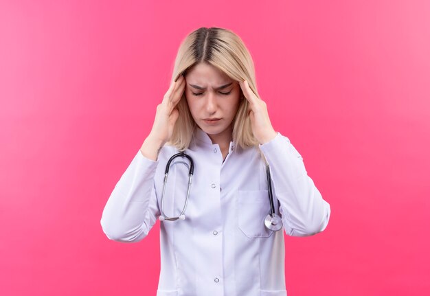 Das müde blonde junge Mädchen des müden Arztes, das Stethoskop im medizinischen Kleid trägt, legte ihre Hände auf Stirn auf isolierte rosa Wand