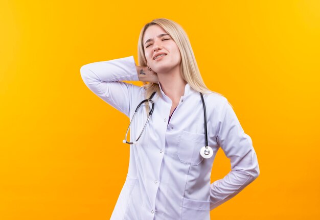 Das müde Mädchen des müden Arztes, das Stethoskop im medizinischen Kleid trägt, legte ihre Hand auf Hals auf isolierte gelbe Wand