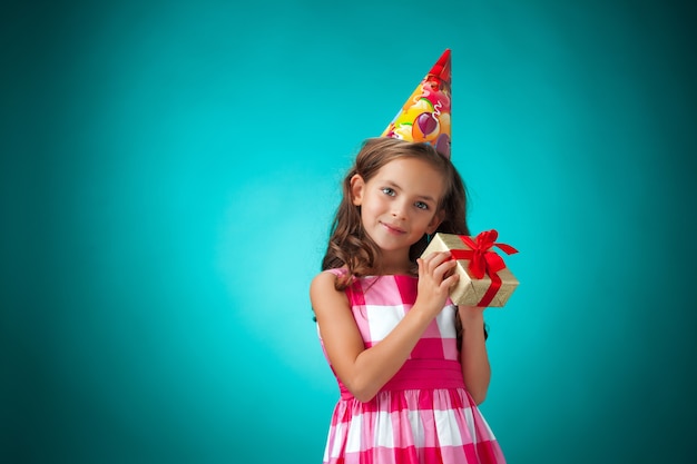 Das niedliche fröhliche kleine Mädchen mit Geschenk und festlicher Kappe auf blauem Hintergrund