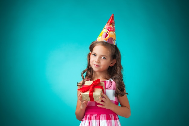 Das niedliche fröhliche kleine Mädchen mit Geschenk und festlicher Kappe auf blauem Hintergrund