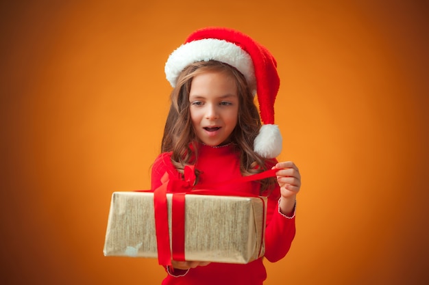 Das niedliche fröhliche kleine Mädchen mit Weihnachtsmütze und Geschenk auf orange Hintergrund