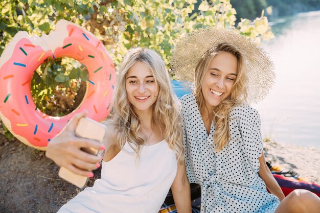 Das Paar der jungen Lesben, das Spaß am Flussufer im sonnigen Tag hat. Frauen verbringen gemeinsam Zeit mit der Natur. Wein trinken, Selfie machen. Konzept der Beziehung, Liebe, Sommer, Wochenende, Flitterwochen.