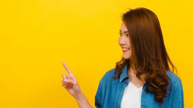 Das Porträt der jungen asiatischen Dame, die mit mit fröhlichem Ausdruck lächelt, zeigt etwas Erstaunliches an der leeren Stelle in der Freizeitkleidung und betrachtet Kamera über gelber Wand. Gesichtsausdruck Konzept.