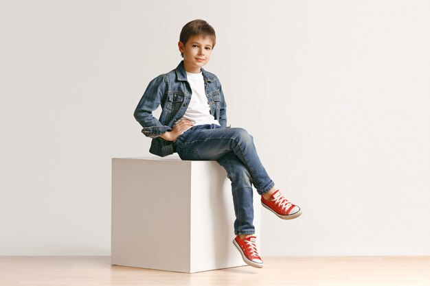 Das Porträt des niedlichen kleinen Jungen in der stilvollen Jeanskleidung, die Kamera gegen weiße Studiowand betrachtet. Kindermode-Konzept
