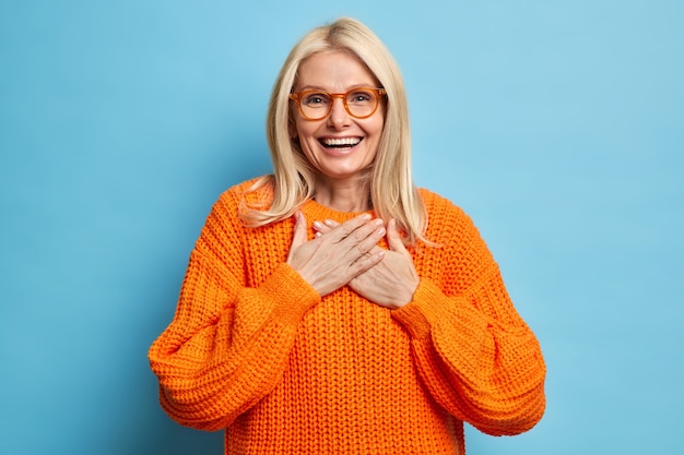 Das Porträt einer blonden Frau mit fröhlichem Ausdruck hält die Hände an die Brust gedrückt und drückt die Dankbarkeit für das herzerwärmende Kompliment aus. Sie trägt eine Brille und einen orangefarbenen Pullover.