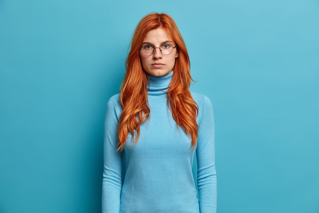 Das Porträt einer ernsthaften, gut aussehenden Ingwerfrau sieht direkt mit den Händen nach unten aus und hat einen selbstbewussten Ausdruck, gekleidet in einen blauen Rollkragenpullover und eine Brille.