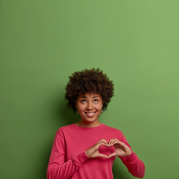 Das Porträt einer fröhlich angenehm aussehenden afroamerikanischen Frau zeigt ein Herzzeichen über der Brust, drückt Liebe und Fürsorge aus, schaut nach oben, trägt einen rosa Pullover, posiert gegen eine grüne Wand, leerer Raum nach oben