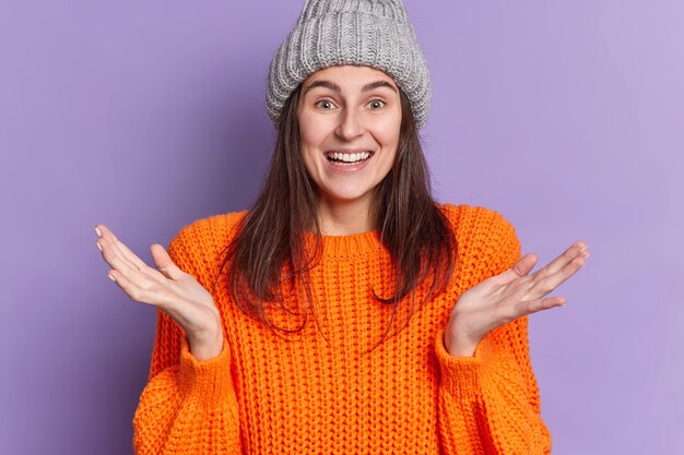 Kostenloses Foto das porträt einer glücklich lächelnden frau hebt die handflächen und lässt die ahnungslose geste mit den schultern zucken. die schultern tragen einen winterorangen pullover und der hut hat dunkles haar.