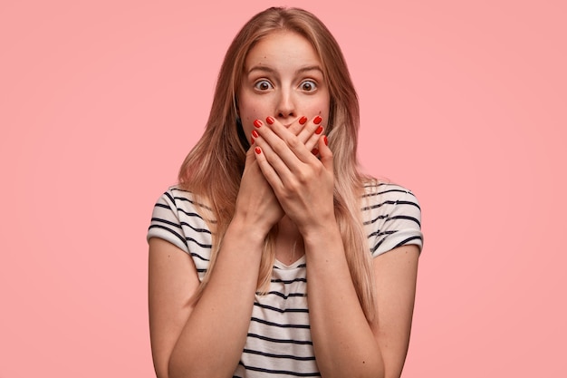 Kostenloses Foto das porträt einer gut aussehenden betäubten jungen frau bedeckt den mund mit beiden händen, starrt in die kamera, versucht sprachlos zu sein, steht drinnen an der rosa wand