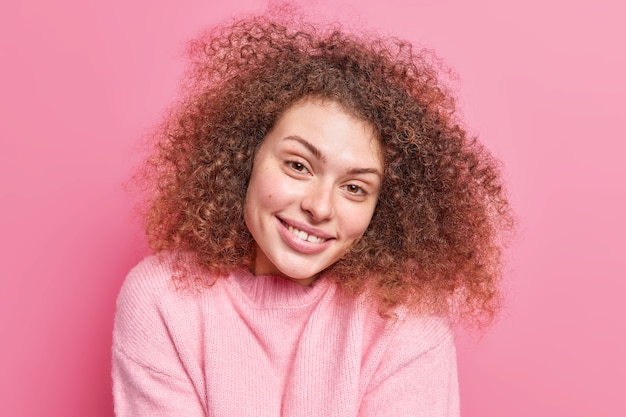 Das Porträt einer positiven verführerischen Frau mit lockigem, buschigem Haar lächelt sanft und zeigt, dass weiße Zähne gesunde Haut haben und einen lässigen Pullover einzeln über einer rosafarbenen Wand tragen. Natürliches Schönheitskonzept