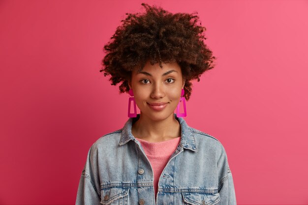 Das Porträt einer schönen afroamerikanischen Frau hat einen charmanten Ausdruck, eine dunkle Haut, trägt Ohrringe und eine Jeansjacke, sieht zufrieden aus und ist bereit, mit Freunden auszugehen