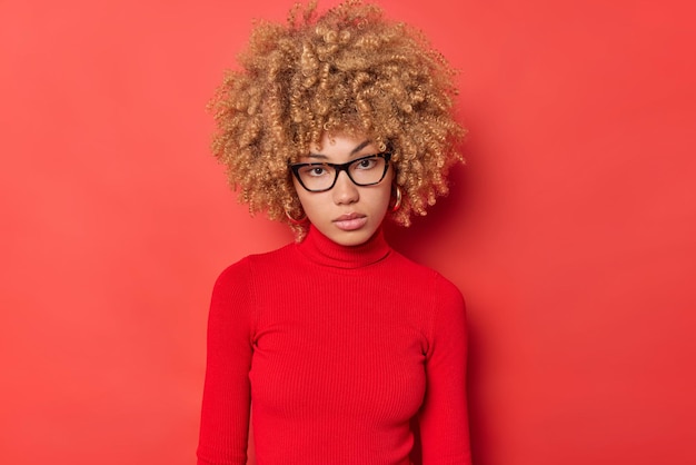 Das Porträt einer seriösen Studentin mit lockigem Haar fühlt sich selbstbewusst an, blickt direkt in die Kamera, trägt eine transparente Brille und einen Rollkragenpullover, der isoliert vor leuchtend rotem Hintergrund steht, und hört Ihnen aufmerksam zu