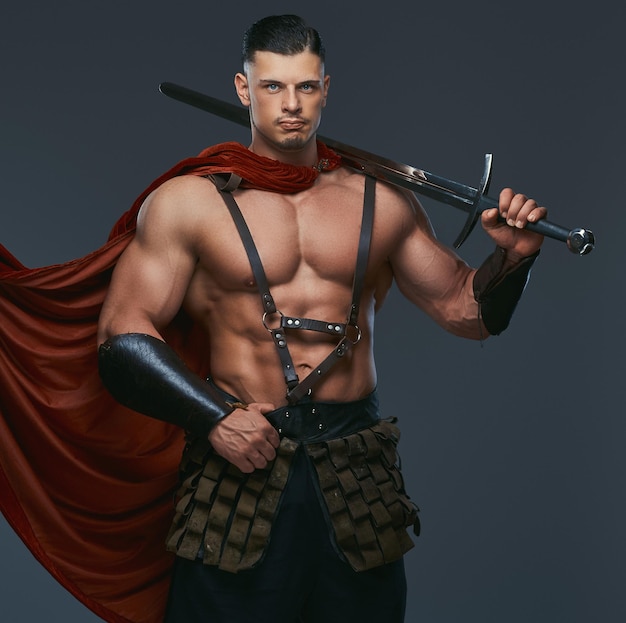 Das Porträt eines antiken griechischen Kriegers mit einem muskulösen Körper, der in Kampfuniformen gekleidet ist, hält ein Schwert auf seiner Schulter. Isoliert auf grauem Hintergrund.