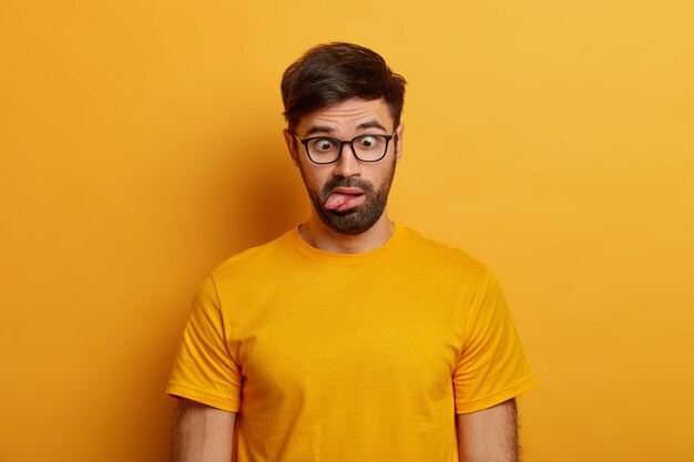 Das Porträt eines bärtigen Mannes zeigt eine Grimasse, kreuzt die Augen und streckt die Zunge heraus, spielt herum, wird verrückt, trägt eine Brille, ein alltägliches T-Shirt und posiert an der gelben Wand. Menschliche Gesichtsausdrücke