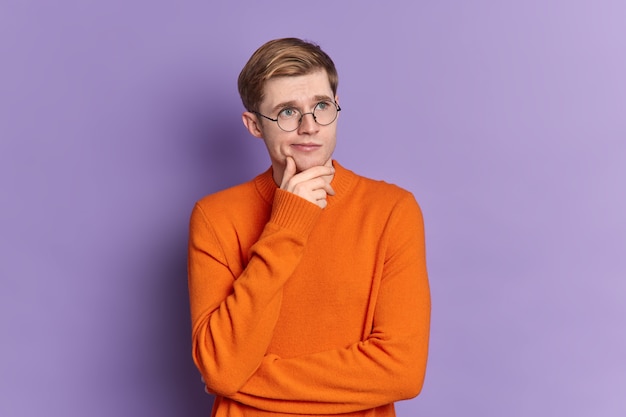 Das Porträt eines blauäugigen Mannes, der über etwas nachdenkt, das in nachdenklicher Haltung steht, hält das Kinn in der Ferne konzentriert und trägt einen lässigen orangefarbenen Pullover