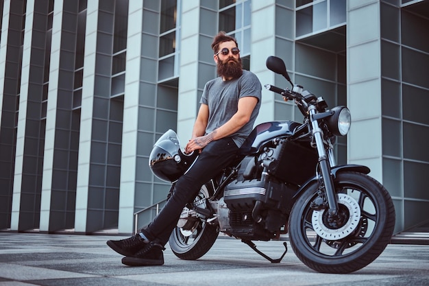 Das Porträt eines brutalen bärtigen Mannes in einem grauen T-Shirt und einer schwarzen Hose hält einen Helm, der auf seinem maßgeschneiderten Retro-Motorrad gegen einen Wolkenkratzer sitzt.