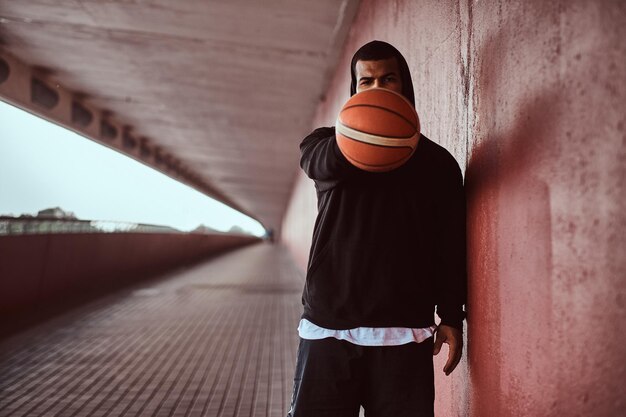 Das Porträt eines dunkelhäutigen Mannes in einem schwarzen Hoodie und Sportshorts hält einen Basketball, während er auf einem Fußweg unter einer Brücke steht.
