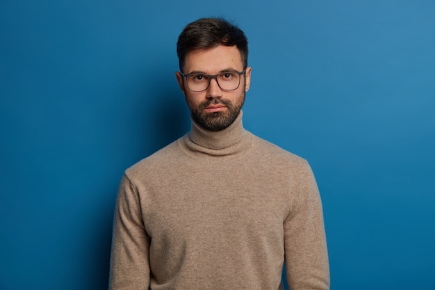 Kostenloses Foto das porträt eines ernsthaften gutaussehenden mannes hat dunkles haar, dicke borsten, schaut direkt in die kamera, trägt eine optische brille und einen rollkragenpullover, isoliert über blauem hintergrund