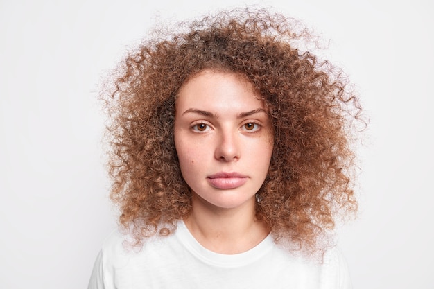 Das Porträt eines ernsthaften tausendjährigen Mädchens hat gesunde Haut, braunes, lockiges, buschiges Haar, das lässig gekleidet aussieht, mit ruhigem Gesichtsausdruck, das ihre natürliche Schönheit isoliert auf weißer Wand zeigt